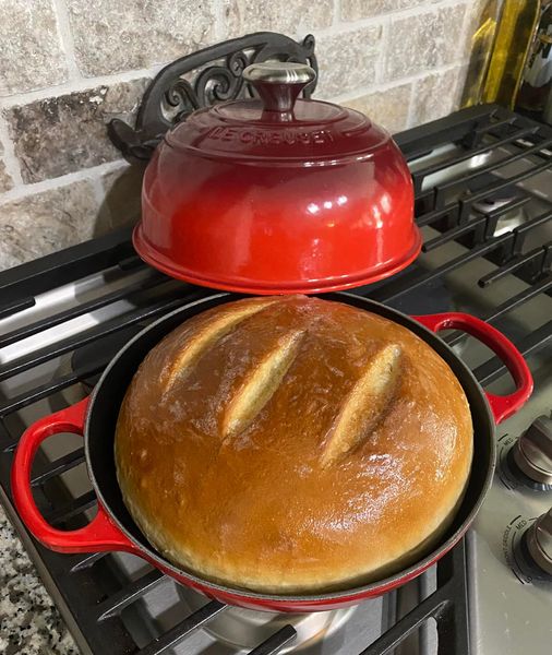 A golden round loaf of French bread baked in a red cast iron bread oven on a stovetop.