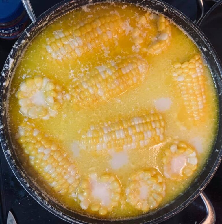 Fresh ears of corn boiling in a rich, opaque yellow broth of melted butter, milk, and sugar inside a large black pot.