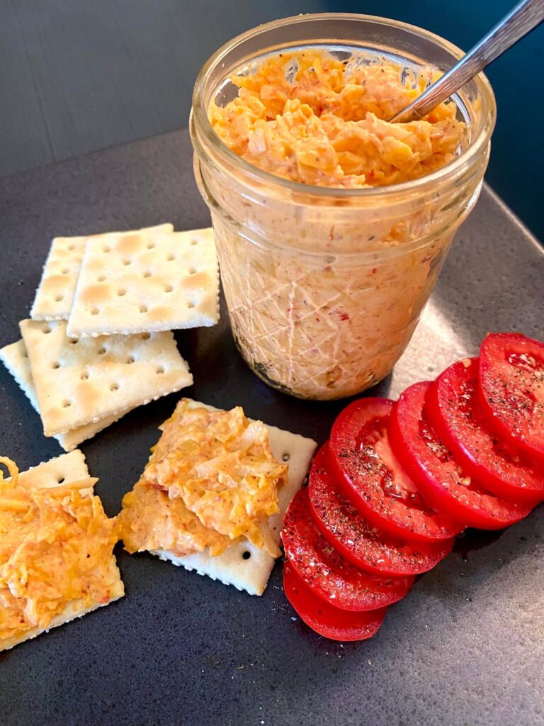 A glass jar filled with creamy southern pimento cheese spread served alongside salted saltine crackers and fresh tomato slices seasoned with pepper.