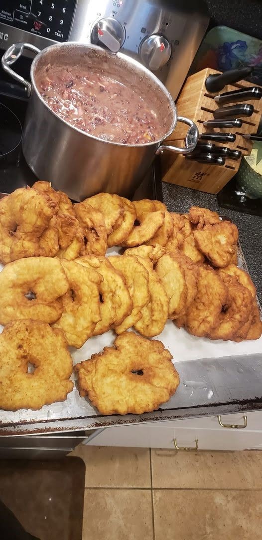 A large batch of freshly fried, golden-brown Indian frybread resting on a baking sheet, with a large pot of hearty beans simmering on the stove in the background.