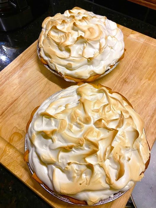 Two deep-dish chocolate meringue pies with tall, golden-brown toasted meringue peaks sitting on a wooden cutting board.