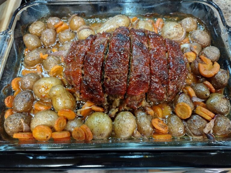 A freshly baked, sliced meatloaf with a shiny red ketchup glaze sitting in the center of a glass baking dish, surrounded by roasted baby potatoes, sliced carrots, and onions in savory pan juices.
