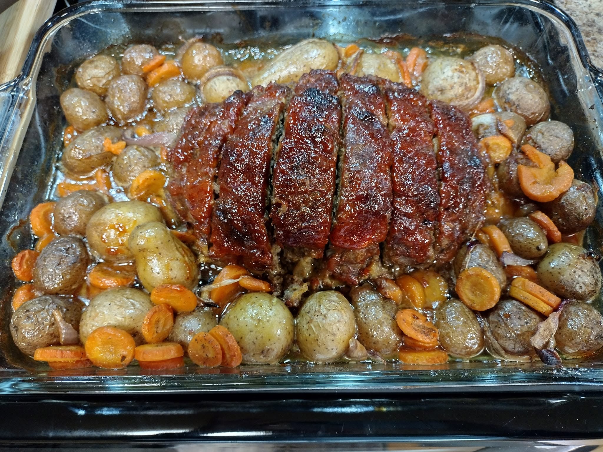 A freshly baked, sliced meatloaf with a shiny red ketchup glaze sitting in the center of a glass baking dish, surrounded by roasted baby potatoes, sliced carrots, and onions in savory pan juices.