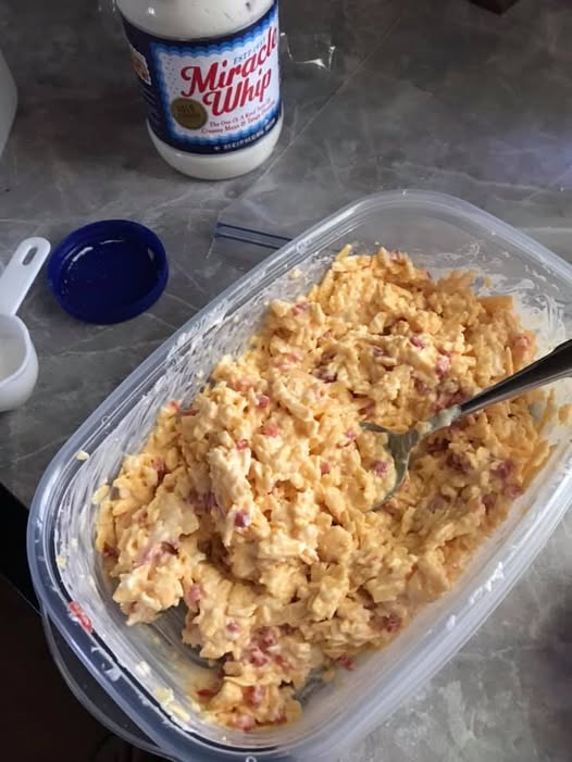 A large clear container filled with freshly mixed, creamy yellow pimiento cheese, with a spoon resting inside and a jar of Miracle Whip in the background.