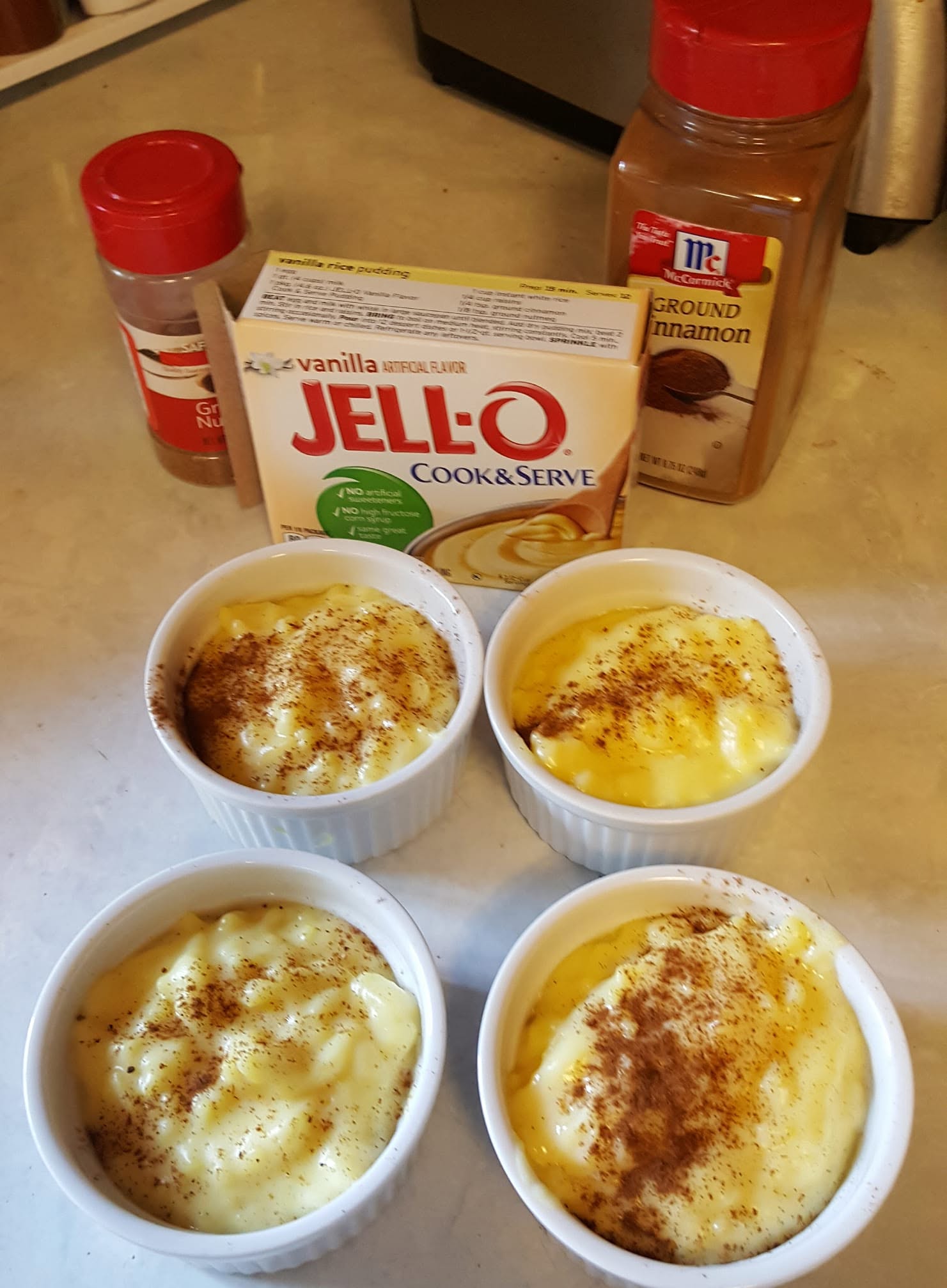 Four white ramekins filled with creamy vanilla rice pudding topped with sprinkled cinnamon and nutmeg, placed next to a box of Jell-O Cook & Serve pudding and spice containers.