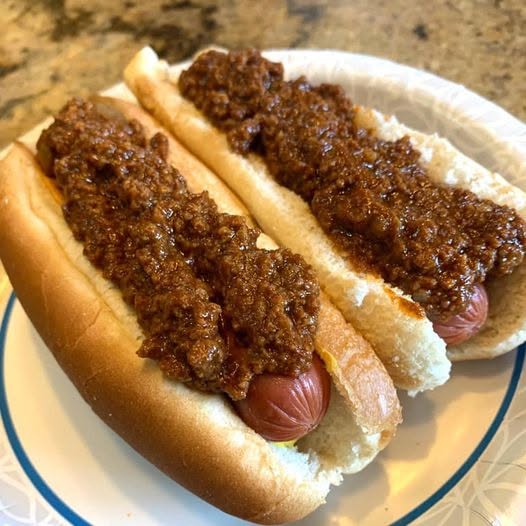 Two classic hot dogs in soft buns, generously topped with a thick, rich, finely textured homemade meat chili sauce, served on a paper plate.