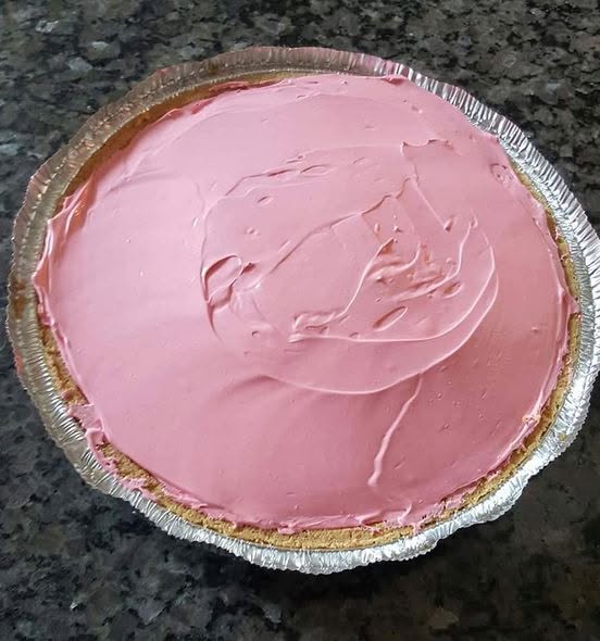 A whole, bright pink no-bake Kool-Aid pie in a graham cracker crust sitting on a dark countertop.
