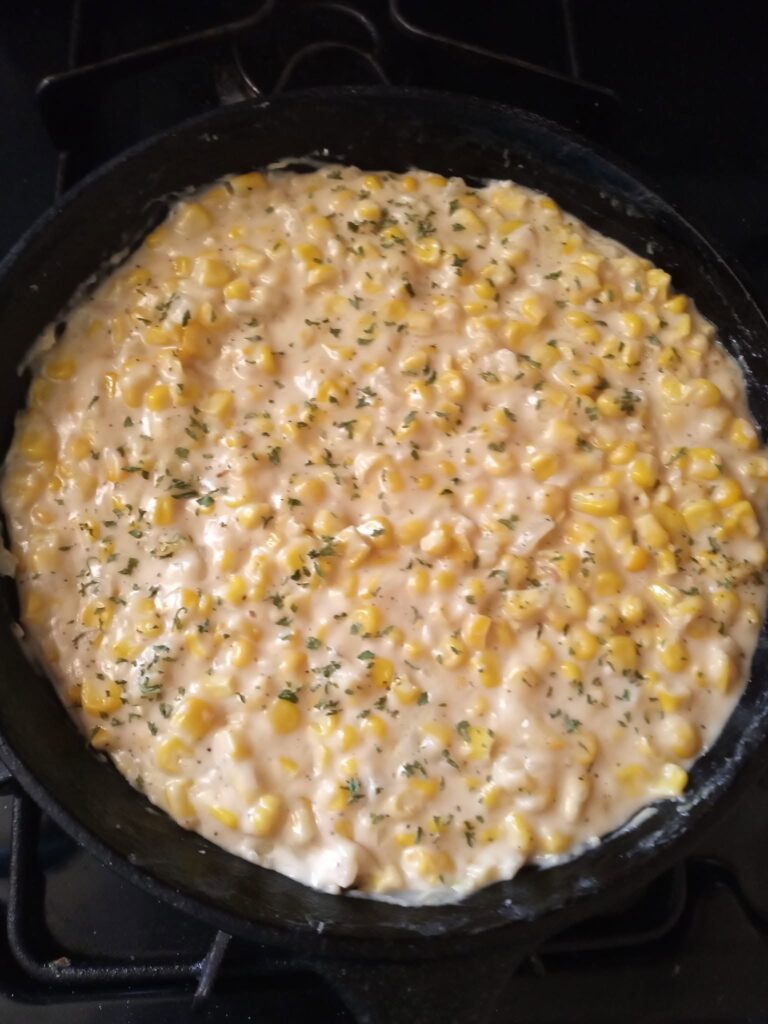 A heavy black cast-iron skillet filled with rich, creamy homemade creamed corn dotted with fresh sweet corn kernels and topped with herbs.
