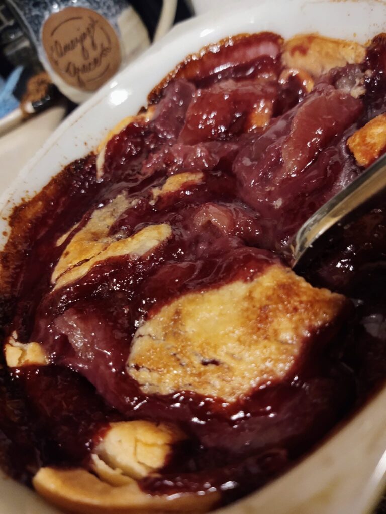 A freshly baked, bubbling blackberry cobbler in a white casserole dish, showing thick purple fruit filling and golden-brown baked pie crust dumplings.