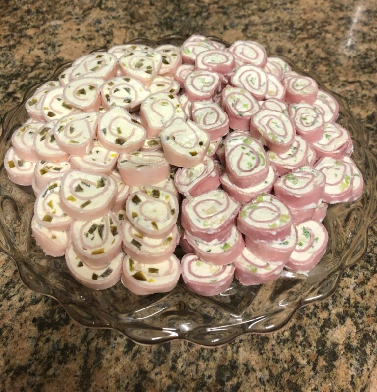 A glass platter piled high with bite-sized sliced ham and turkey roll-ups filled with a creamy green onion and jalapeño cream cheese spread.