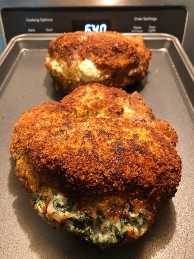 Two thick, golden-brown breaded pork chops stuffed with a creamy spinach and cheese filling, resting on a metal baking sheet.