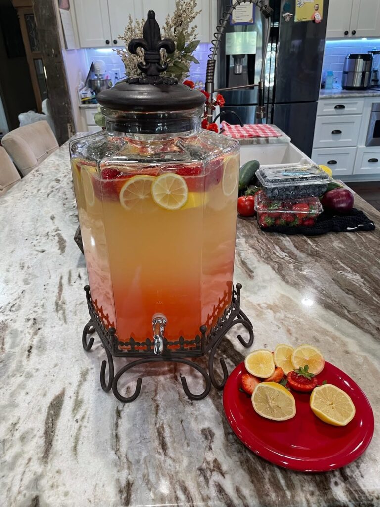 A large glass beverage dispenser filled with layered fresh strawberry lemonade, sitting on a kitchen counter next to a red plate of sliced lemons and strawberries.