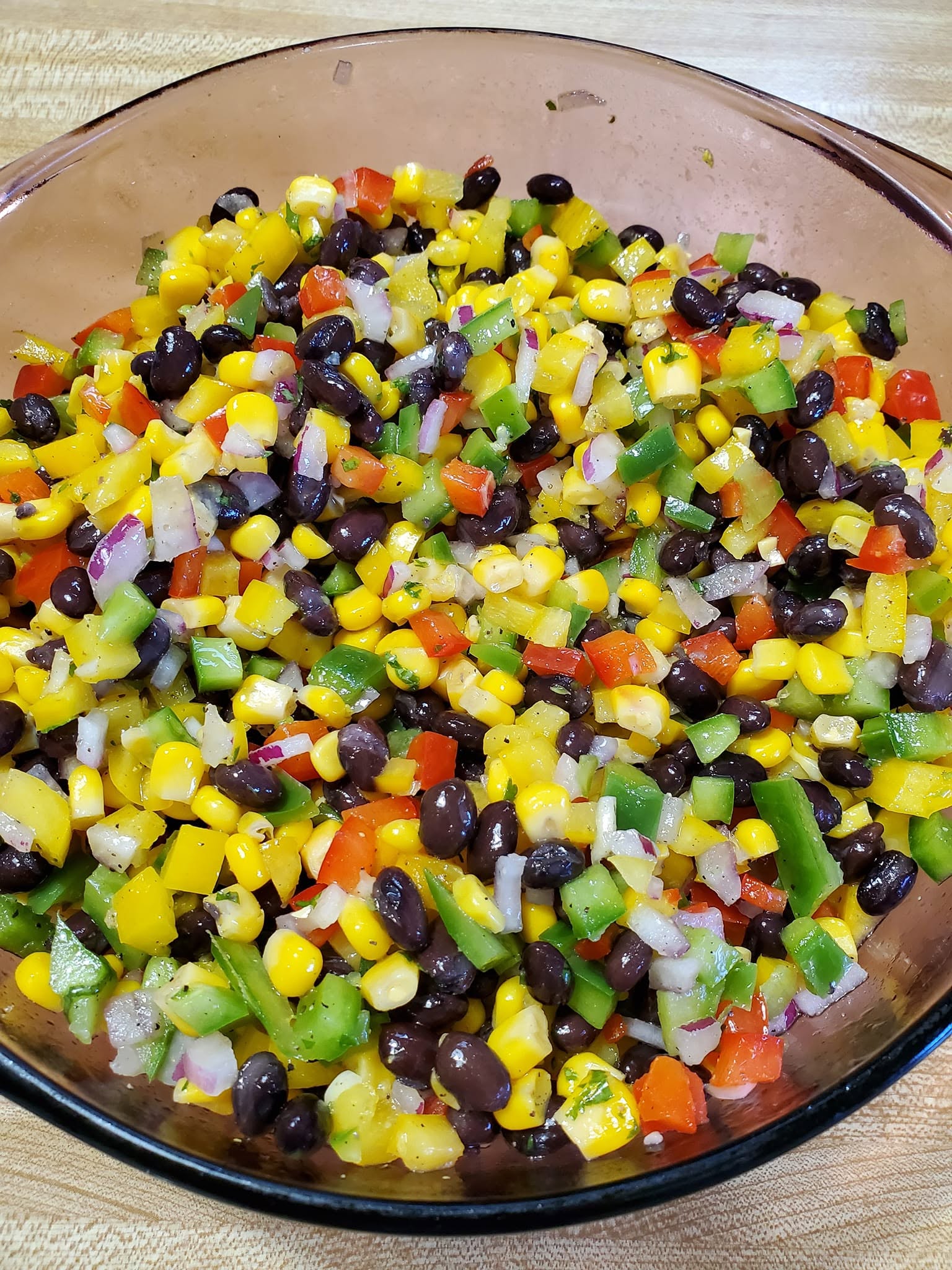 A large glass bowl filled with a cold, colorful summer black bean and corn salad featuring diced red, green, and yellow bell peppers and black beans.