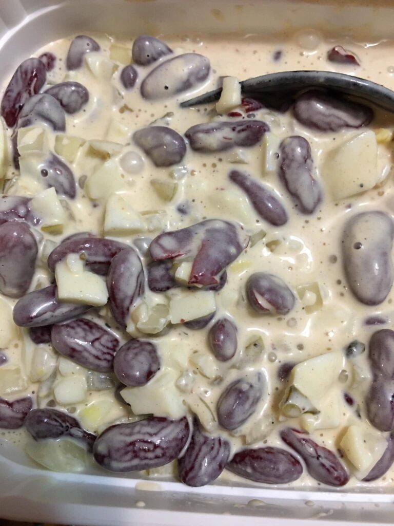 A close-up view of a large container filled with a vintage-style creamy kidney bean salad, featuring dark red kidney beans and diced hard-boiled eggs coated in a tangy dressing.