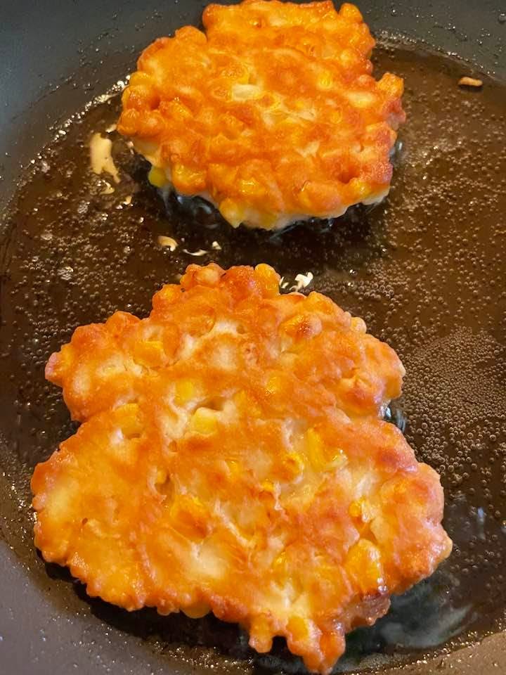 Two golden-brown corn fritters frying in a skillet of hot oil with visible bubbles around the edges.