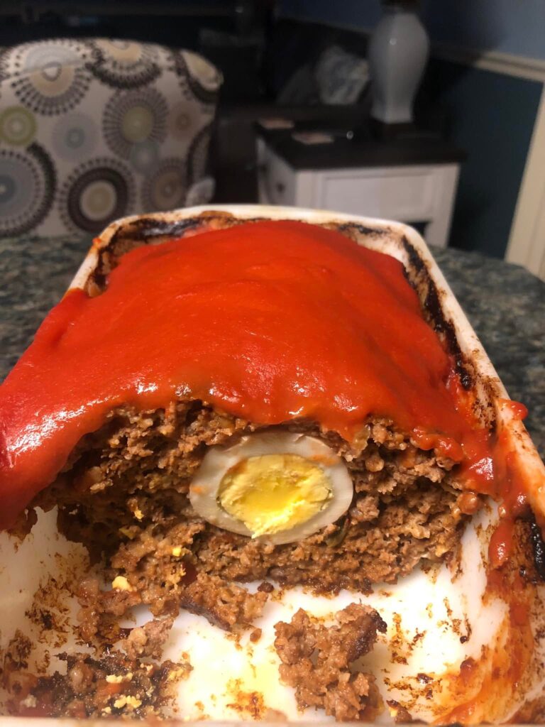 A sliced view of a baked meatloaf topped with a thick red glaze, revealing a perfectly cooked hard-boiled egg hidden in the center of the savory beef.
