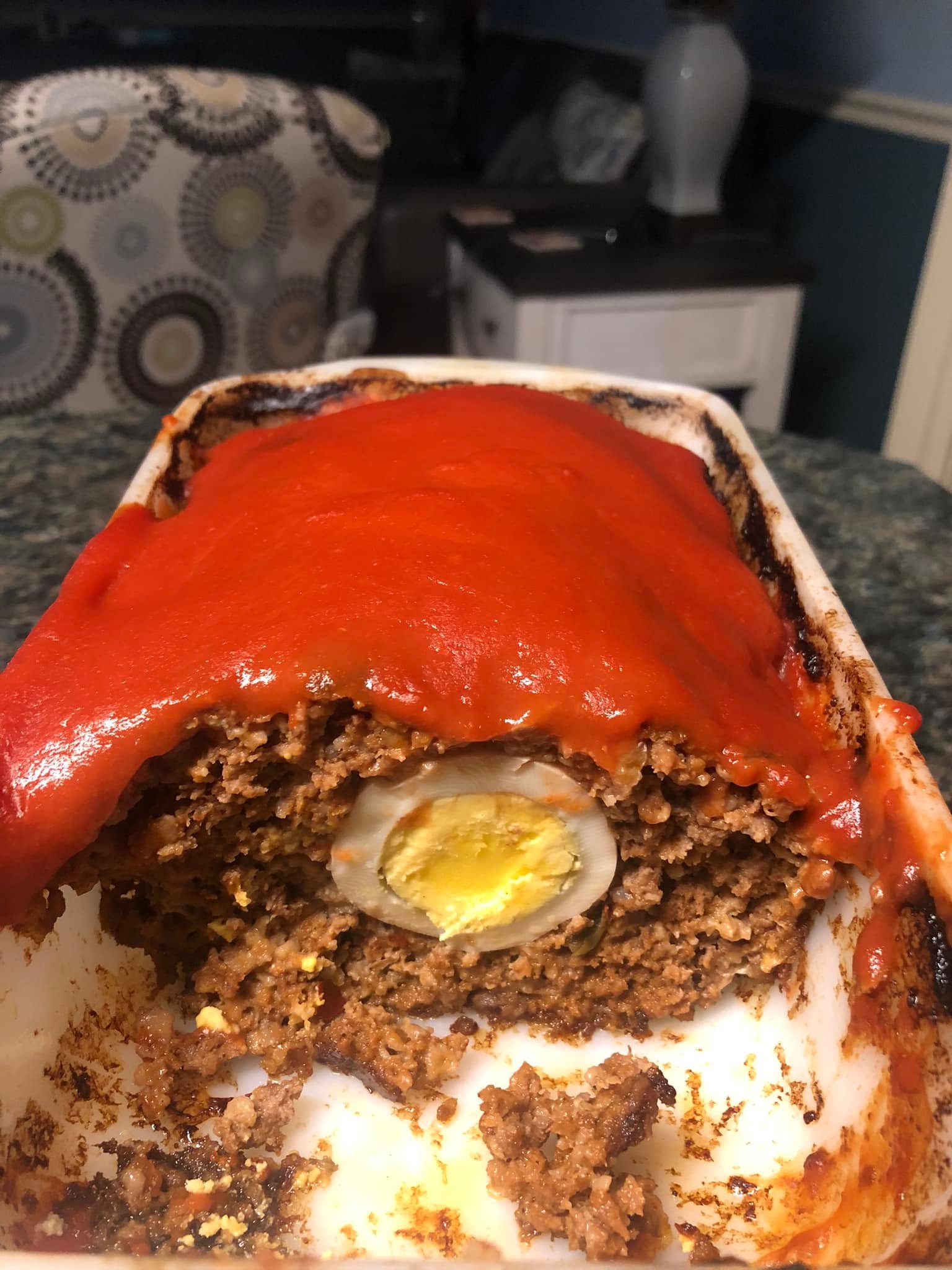 A sliced view of a baked meatloaf topped with a thick red glaze, revealing a perfectly cooked hard-boiled egg hidden in the center of the savory beef.