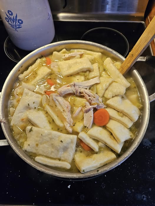 A large stovetop pot filled with rich, bubbling chicken broth, shredded chicken, carrots, and soft, pillowy southern-style rolled dumplings.