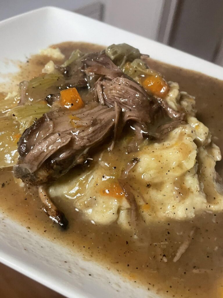 A hearty serving of tender, shredded pot roast with carrots and celery, smothered in savory natural juices over a bed of rustic smashed potatoes heavily seasoned with black pepper.