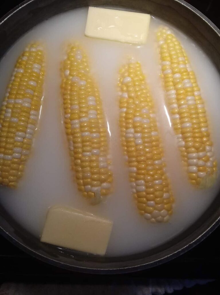 Fresh ears of corn boiling in a pot filled with water, a cup of white milk, a stick of butter, sugar, and salt.