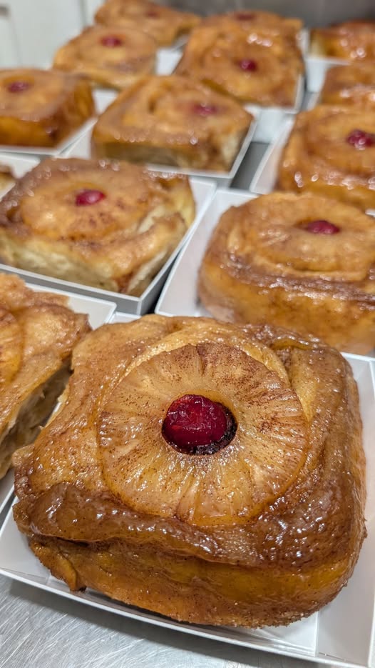 A close up of a freshly baked, glossy pineapple upside down cinnamon roll featuring a caramelized pineapple ring and a bright red cherry in the center, sitting in a white bakery box.