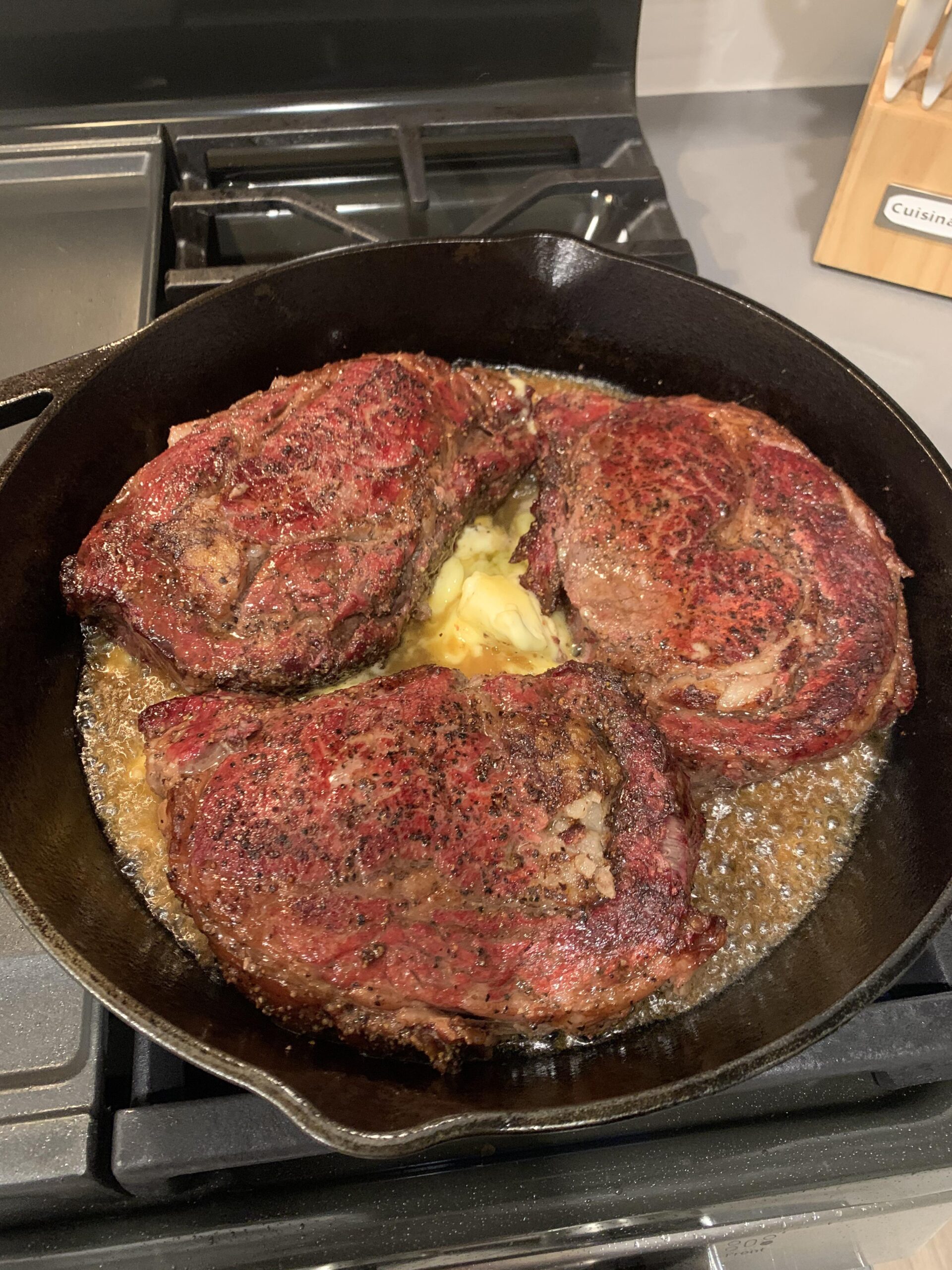 Three perfectly seared, thick-cut ribeye steaks sizzling in a pool of melted butter inside a 12-inch Lodge cast-iron skillet.