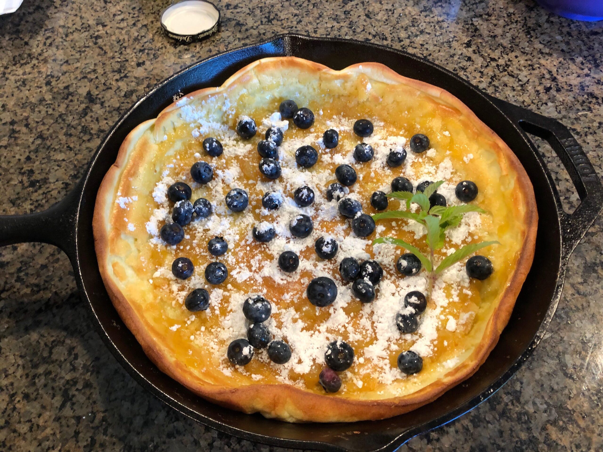 A golden-brown Dutch baby pancake baked in a cast-iron skillet, topped with fresh blueberries, a dusting of powdered sugar, and a sprig of fresh mint.