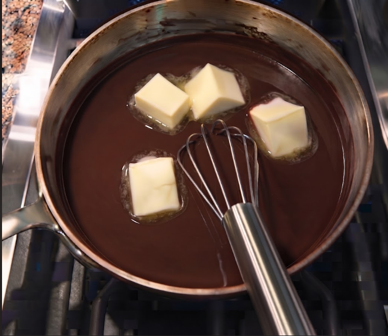 A rich, glossy chocolate fudge icing being poured generously over a freshly baked sheet cake, cascading down the sides.
