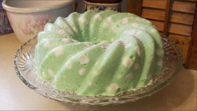A bright green, molded retro jello salad dotted with cottage cheese on a decorative glass serving plate. A Beloved Mid-Century Holiday Dessert