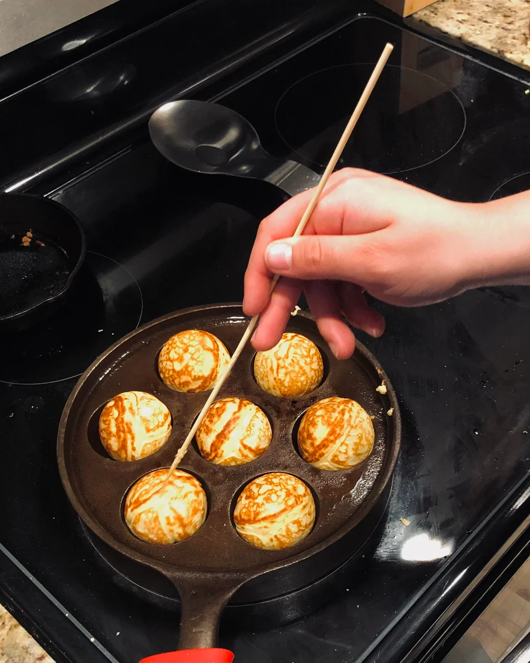 A hand using a wooden skewer to turn golden-brown Aebleskiver pancake balls in a cast-iron Aebleskiver pan on a black stovetop.