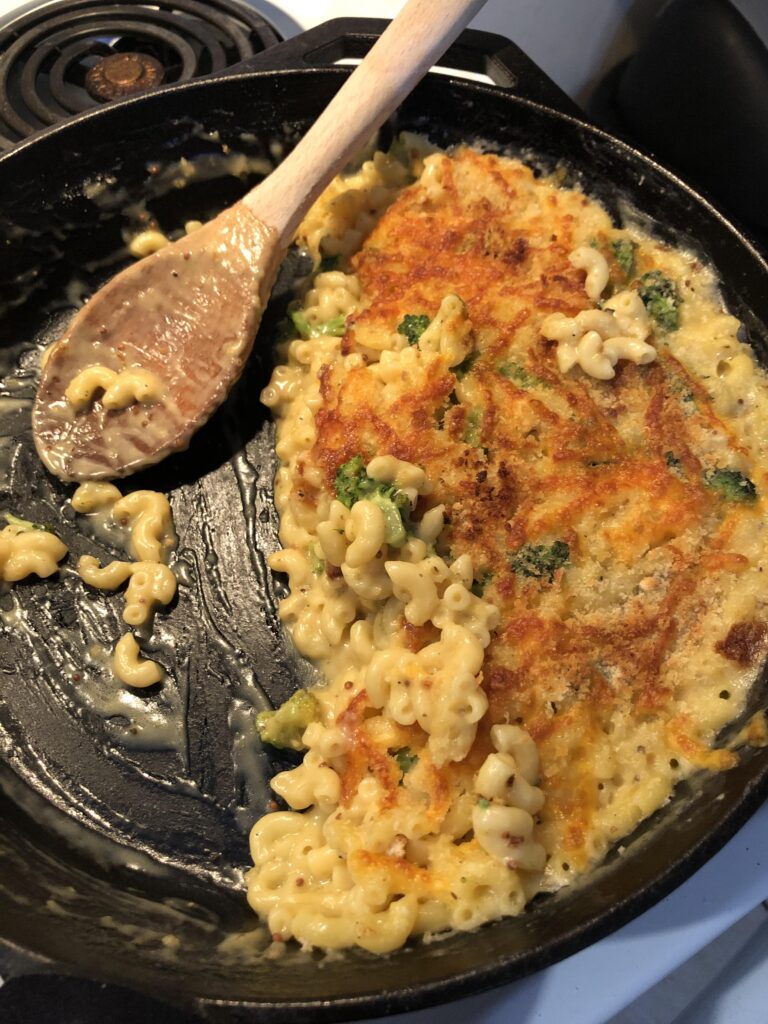 A cast iron skillet filled with creamy mac and cheese mixed with broccoli, topped with a golden brown crispy panko crust, with a wooden spoon resting inside.