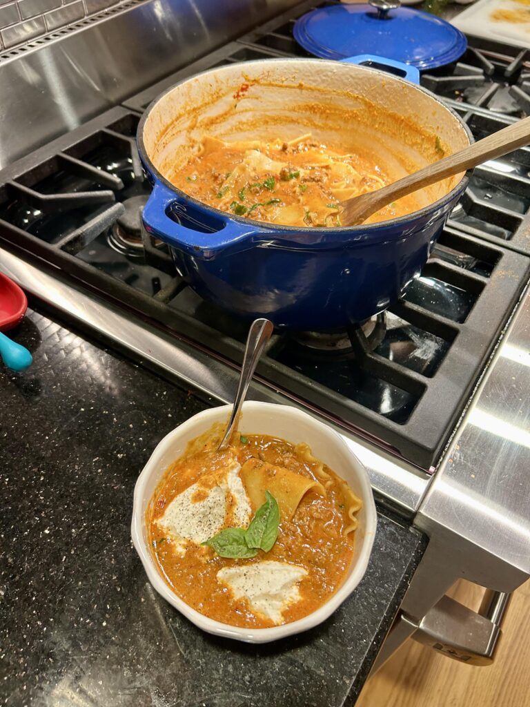 A blue Dutch oven filled with creamy lasagna soup on a stovetop, next to a white bowl of soup topped with seasoned ricotta cheese and fresh basil.