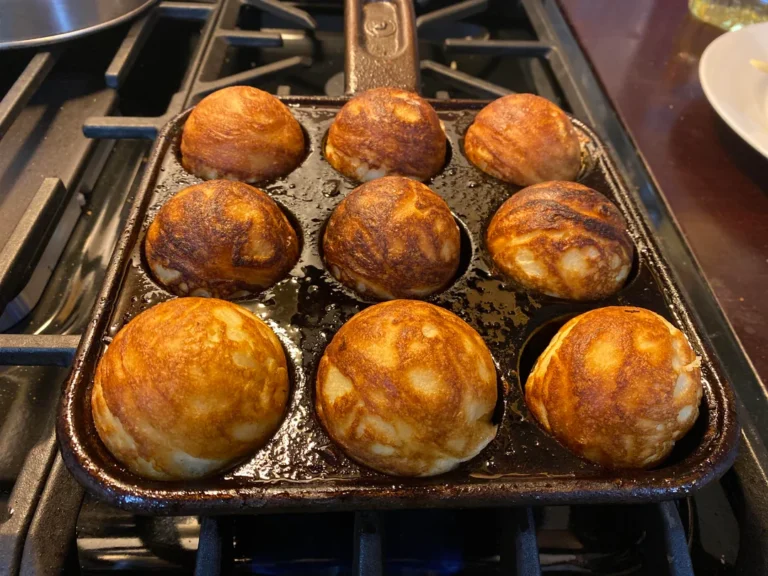 A square cast-iron aebleskiver pan filled with eight perfectly golden-brown, puffed Danish pancake balls cooking on a gas stovetop.