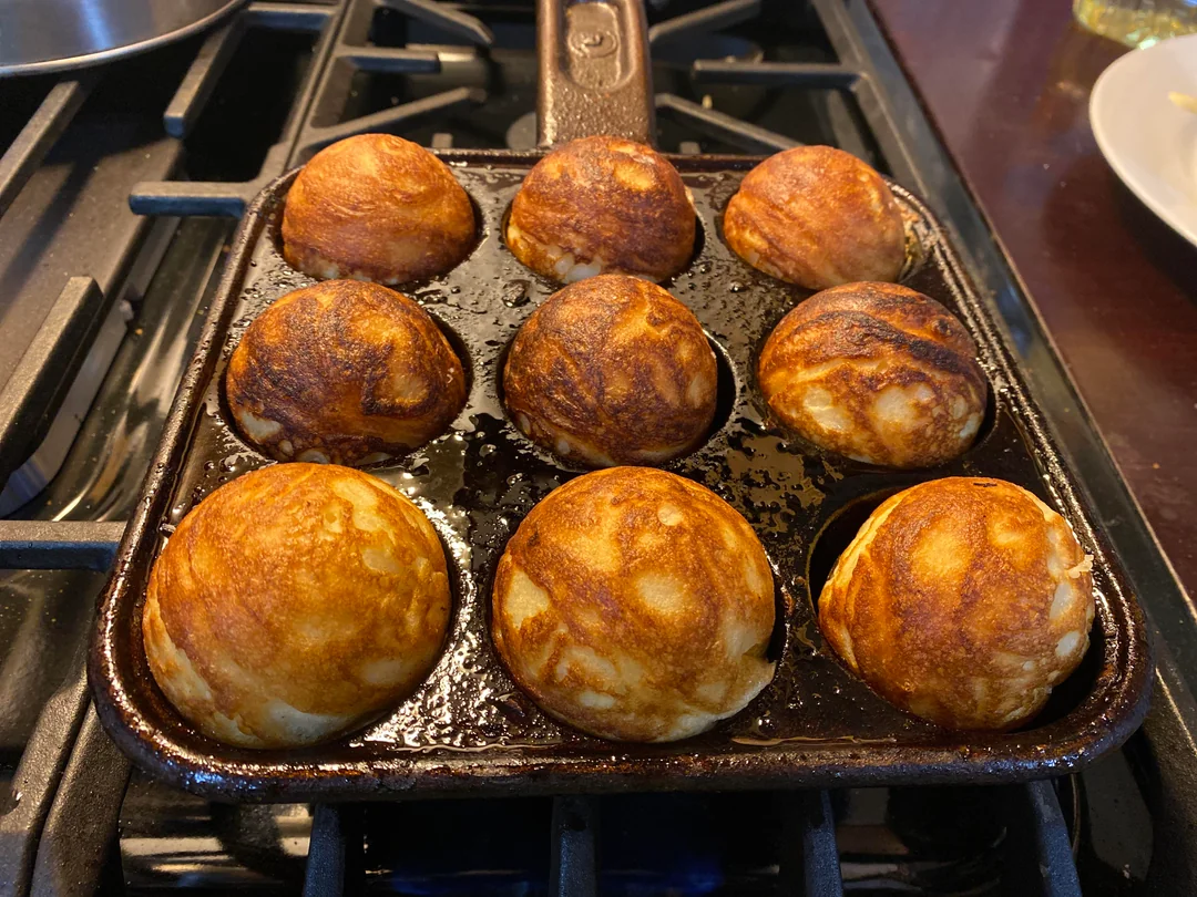 A square cast-iron aebleskiver pan filled with eight perfectly golden-brown, puffed Danish pancake balls cooking on a gas stovetop.