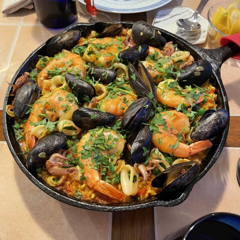 A heavy black cast-iron skillet filled with a vibrant seafood paella, topped with large prawns, mussels, sliced squid, peas, and fresh parsley, served alongside lemon wedges.