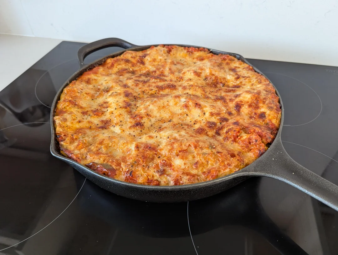 A freshly baked whole cast iron skillet lasagna sitting on a black glass induction stovetop with a golden-brown, bubbly cheese topping.