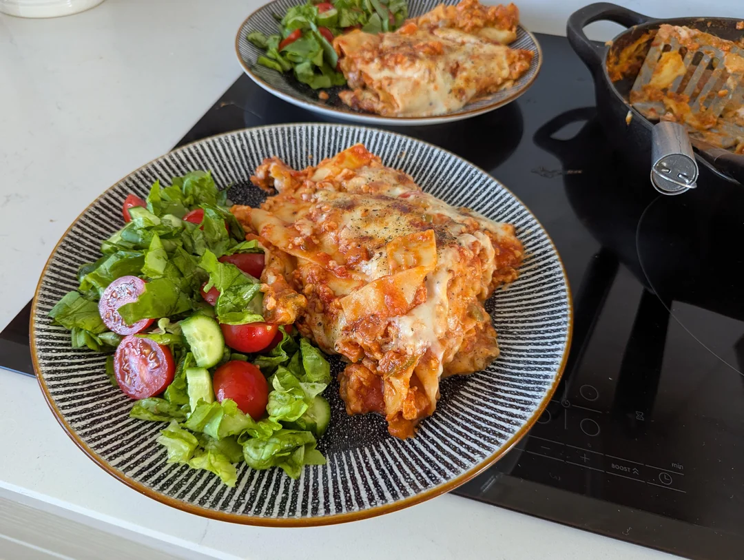 Two plates of freshly served cast iron lasagna alongside a crisp green side salad with cherry tomatoes and cucumbers.