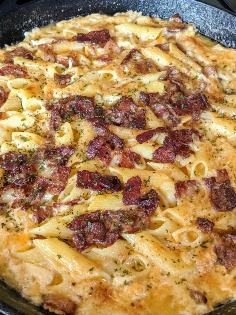 A large cast iron skillet filled with creamy, bubbling baked macaroni and cheese made with penne pasta, topped with crispy bacon pieces and fresh parsley.