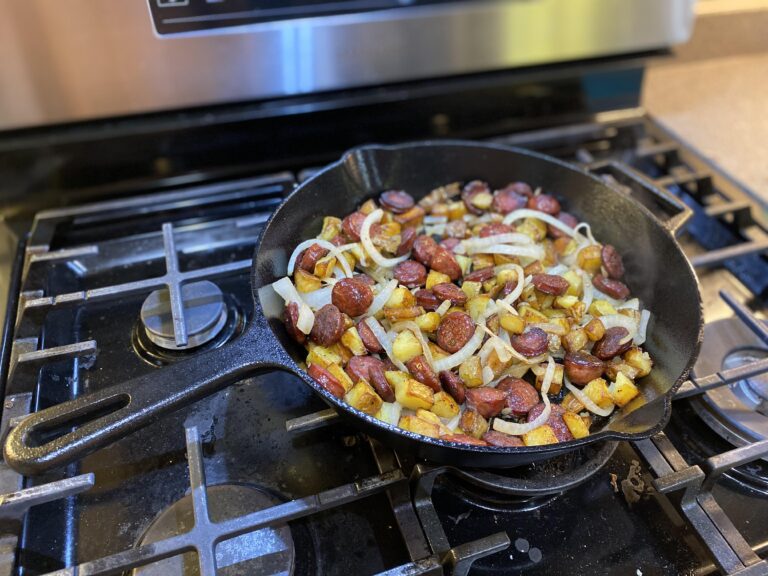 A black cast-iron skillet filled with browned slices of smoked sausage, golden diced potatoes, and tender yellow onions sitting on a gas stove.