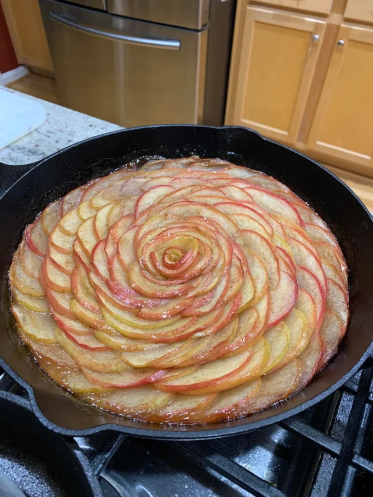 A freshly baked caramel apple rose tart inside a cast-iron skillet, featuring glossy, tender, caramelized apple petals.