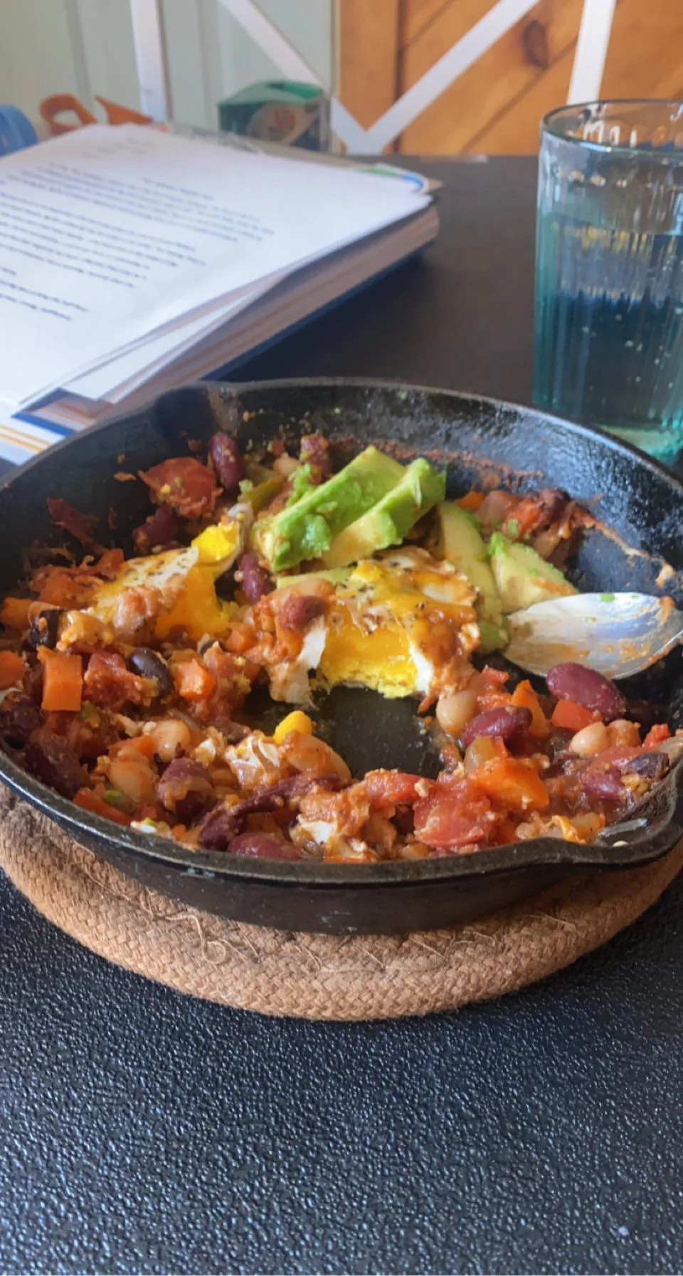 A partially eaten skillet of chili baked eggs showing a perfectly runny, bright yellow yolk mixing with the beans, tomatoes, and fresh avocado.