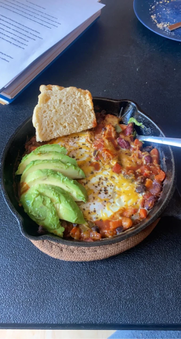 A small cast iron skillet filled with leftover chili, two baked eggs, melted cheddar cheese, and fresh sliced avocado, served with a piece of crusty bread.