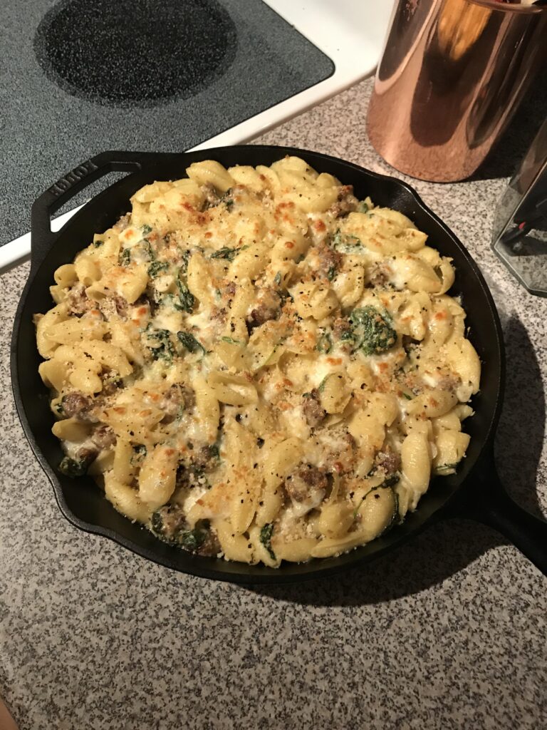 A dark cast iron skillet filled with freshly baked shell pasta, chunks of browned Italian sausage, vibrant green broccoli rabe, and a golden, bubbly cheese crust.