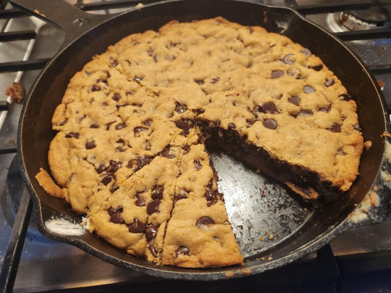 A giant chocolate chip skillet cookie baked inside a black cast iron pan, with a wedge sliced out to reveal a soft, chewy interior.