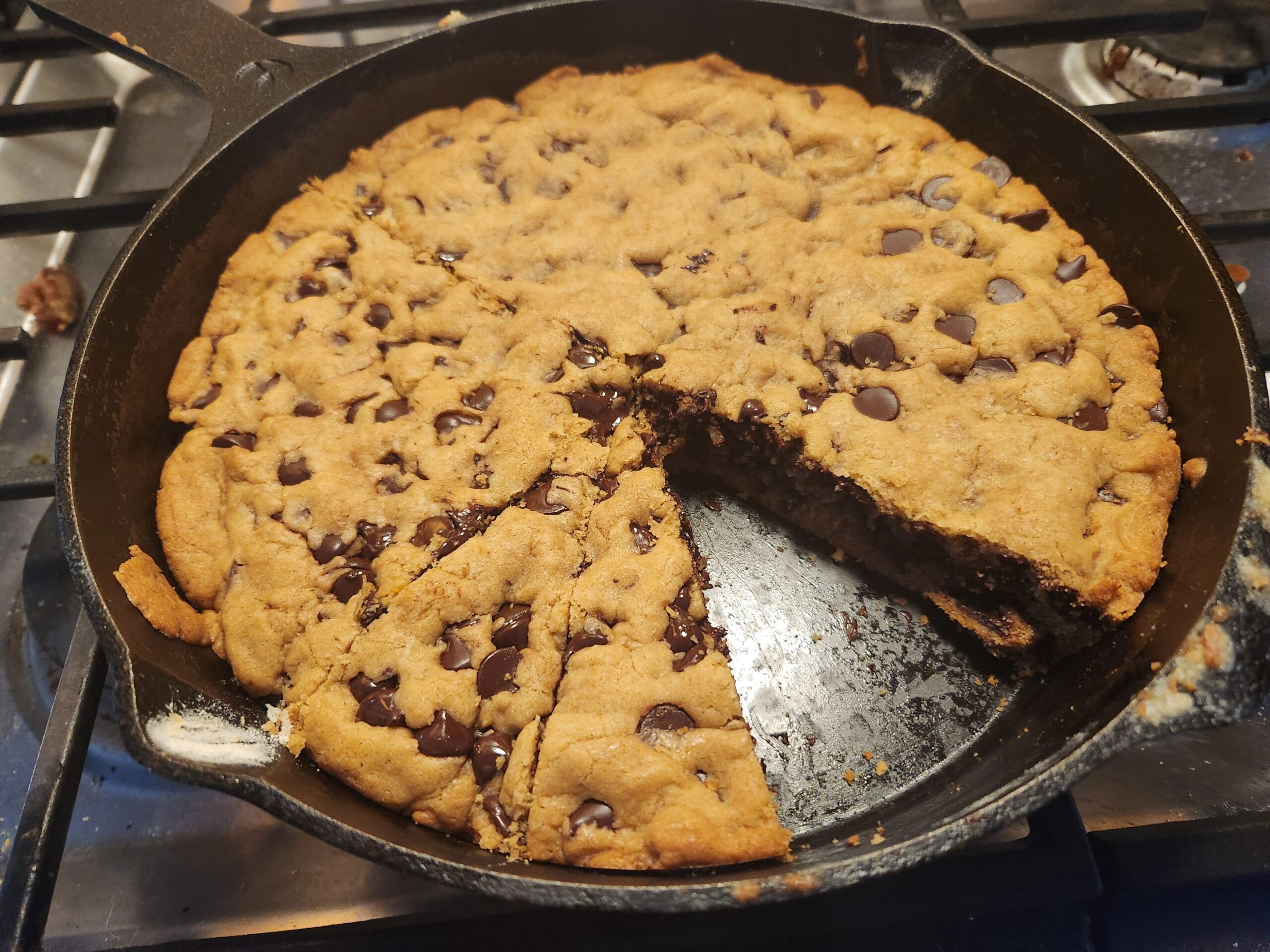 A giant chocolate chip skillet cookie baked inside a black cast iron pan, with a wedge sliced out to reveal a soft, chewy interior.