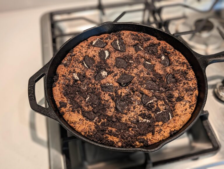 A giant chocolate chip and Oreo cookie baked inside a black cast iron skillet, topped with crunchy cookie pieces and fine Oreo crumbs.