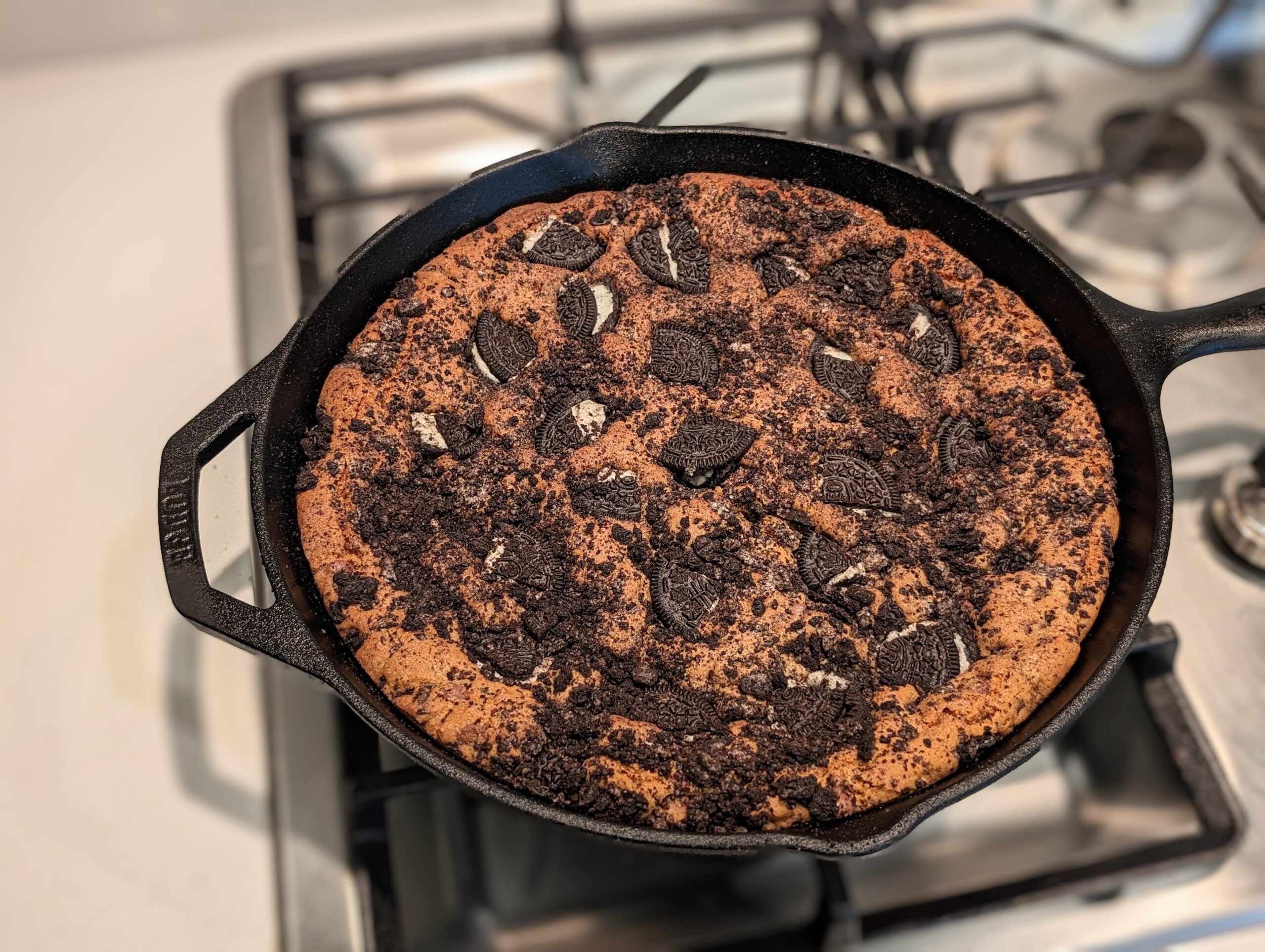 A giant chocolate chip and Oreo cookie baked inside a black cast iron skillet, topped with crunchy cookie pieces and fine Oreo crumbs.