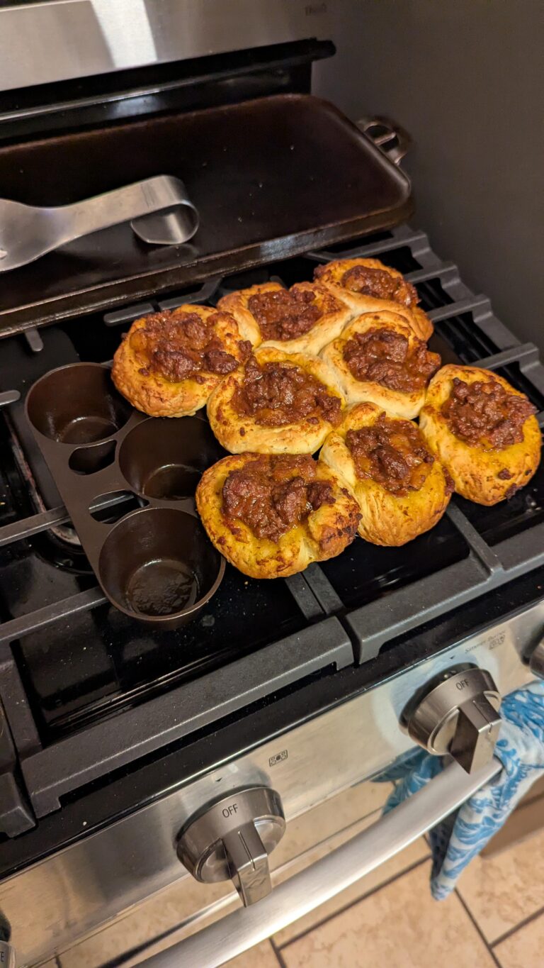 Freshly baked sloppy joe biscuits resting next to a restored vintage Griswold No. 10 muffin pan on a black gas stove.