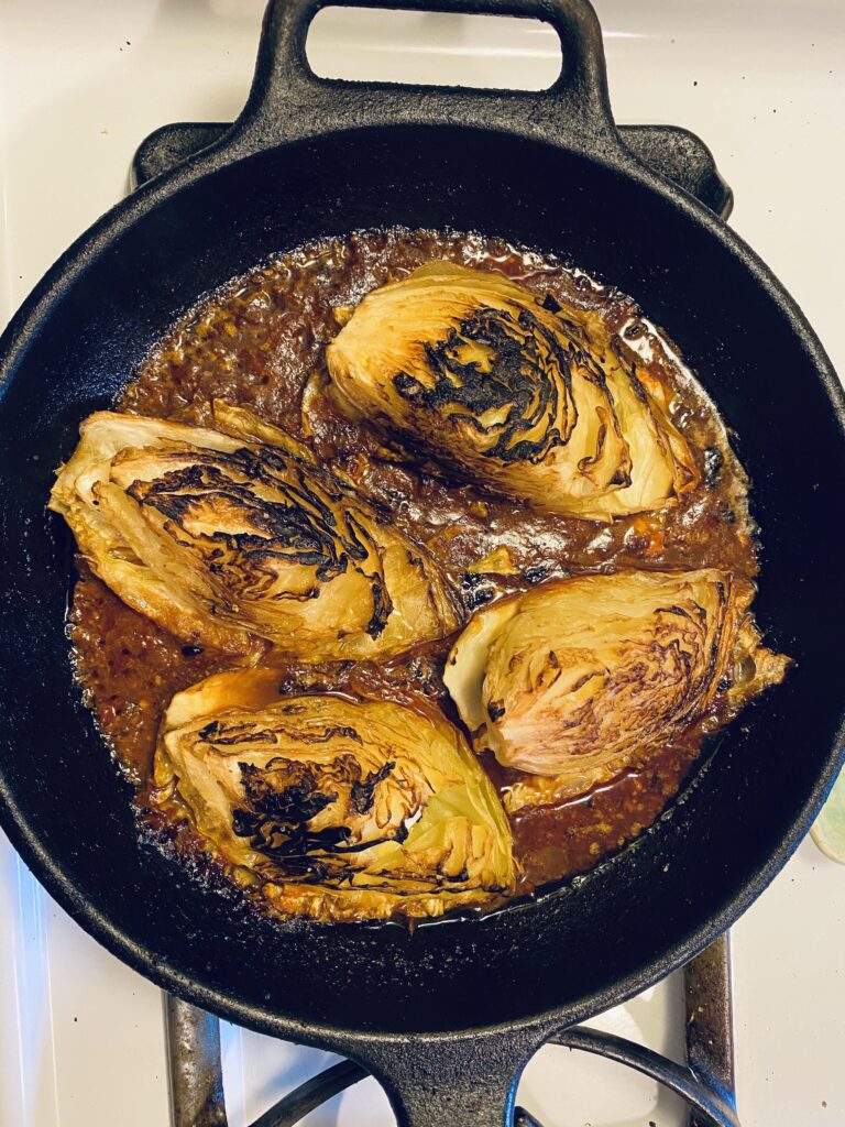 Four wedges of deeply charred, caramelized green cabbage simmering in a rich, savory brown broth inside a large cast iron skillet.