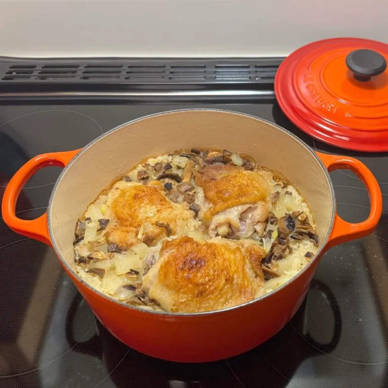 A red Dutch oven filled with golden baked chicken thighs, fluffy long-grain rice, and sautéed mushrooms resting on a glass-top stove.